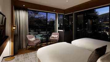 A modern hotel room with a large bed, two armchairs, a small table, and a view of the cityscape.