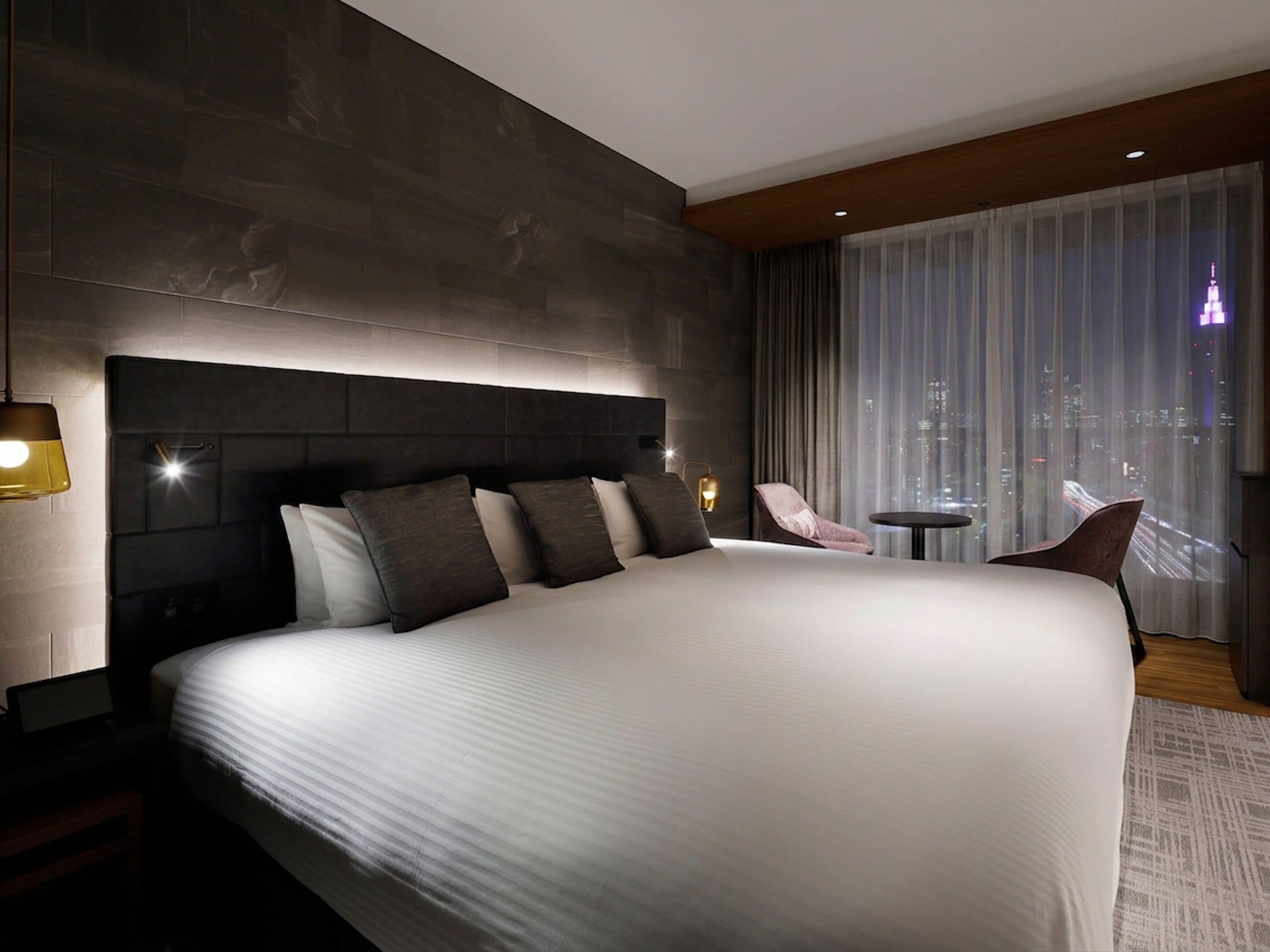 A modern hotel room with a large bed, a nightstand, a chair, a small table, and a city view through the window.