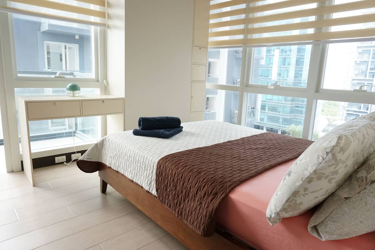A bedroom with a bed, a window, a desk, and a view of buildings.