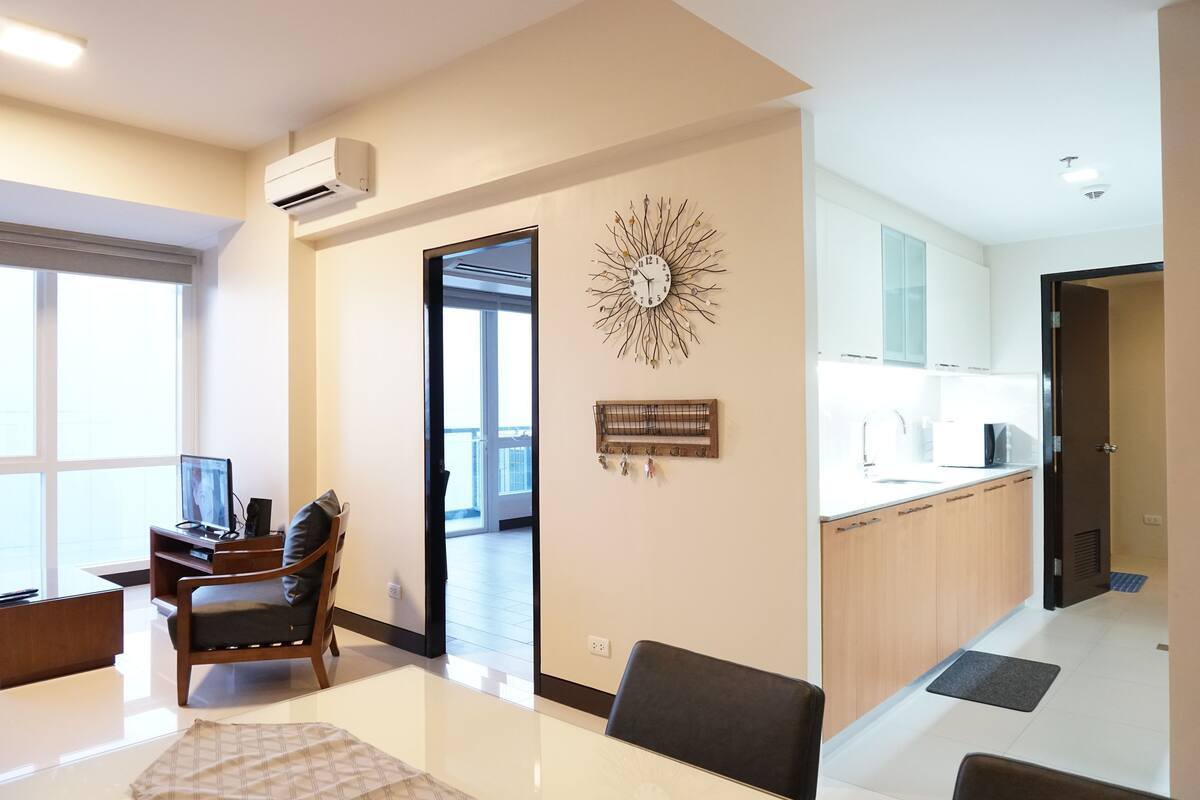 A modern living room with a TV, a sofa, and a dining area. There is a kitchen with cabinets and a sink.