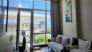 Ein modernes Wohnzimmer mit einer Couch, einem Flachbildfernseher und einem großen Fenster mit Blick auf ein Gebäude und Grünflächen.