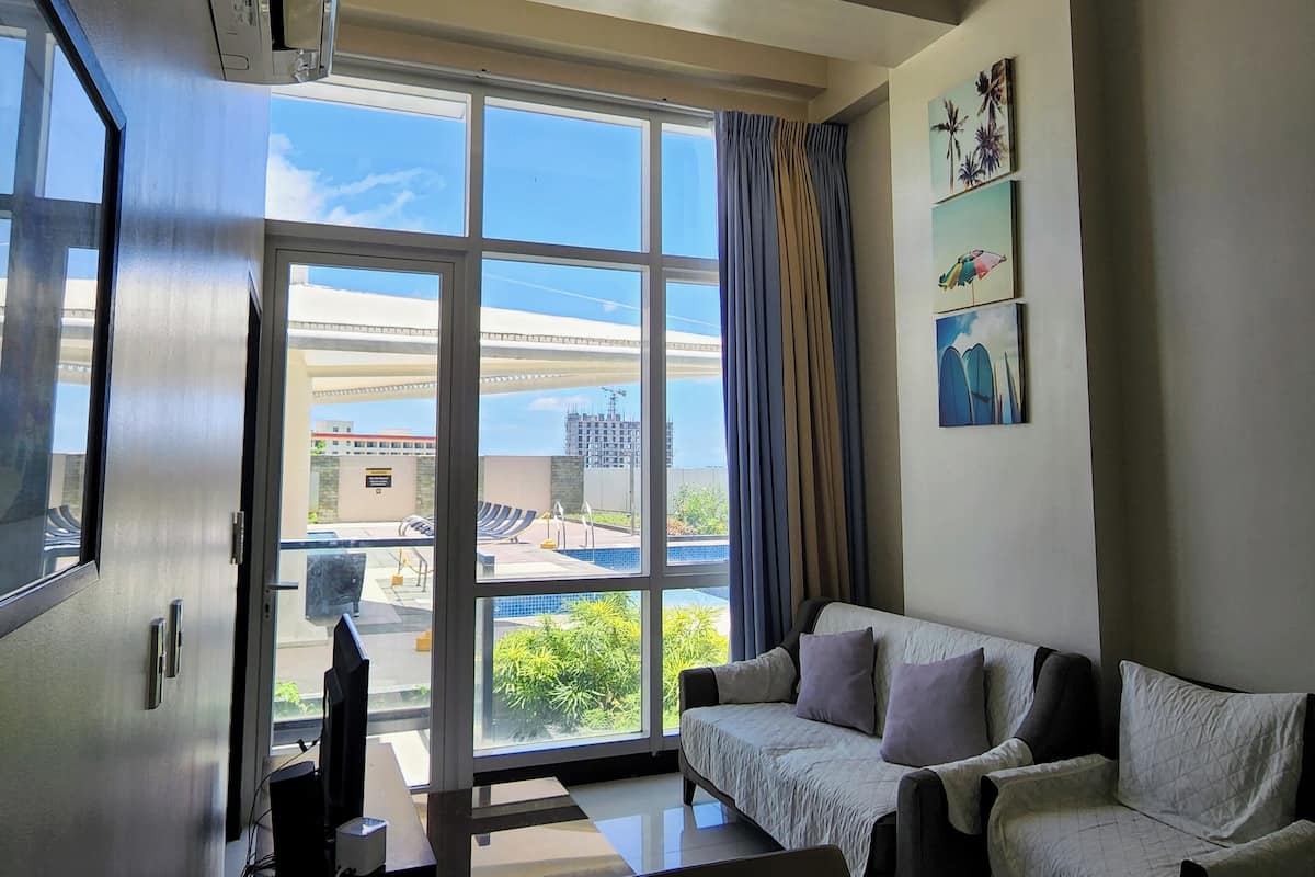 A modern living room with a sofa, a flat-screen TV, and a large window offering a view of a building and greenery.