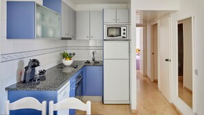 Cocina compacta con armarios azules, un microondas y un refrigerador. Hay una silla blanca y una mesa pequeña con una planta.