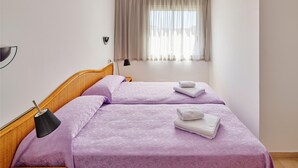 Un dormitorio con dos camas, cada una con una colcha morada y toallas blancas. Hay un cabecero de madera, una lámpara de pie negra y una ventana con cortinas traslúcidas.