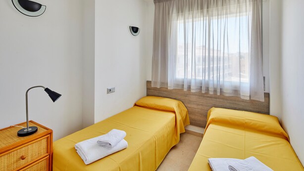 Una habitación de hotel con dos camas individuales, una mesita de noche con lámpara, una ventana con cortinas traslúcidas y vistas a edificios.