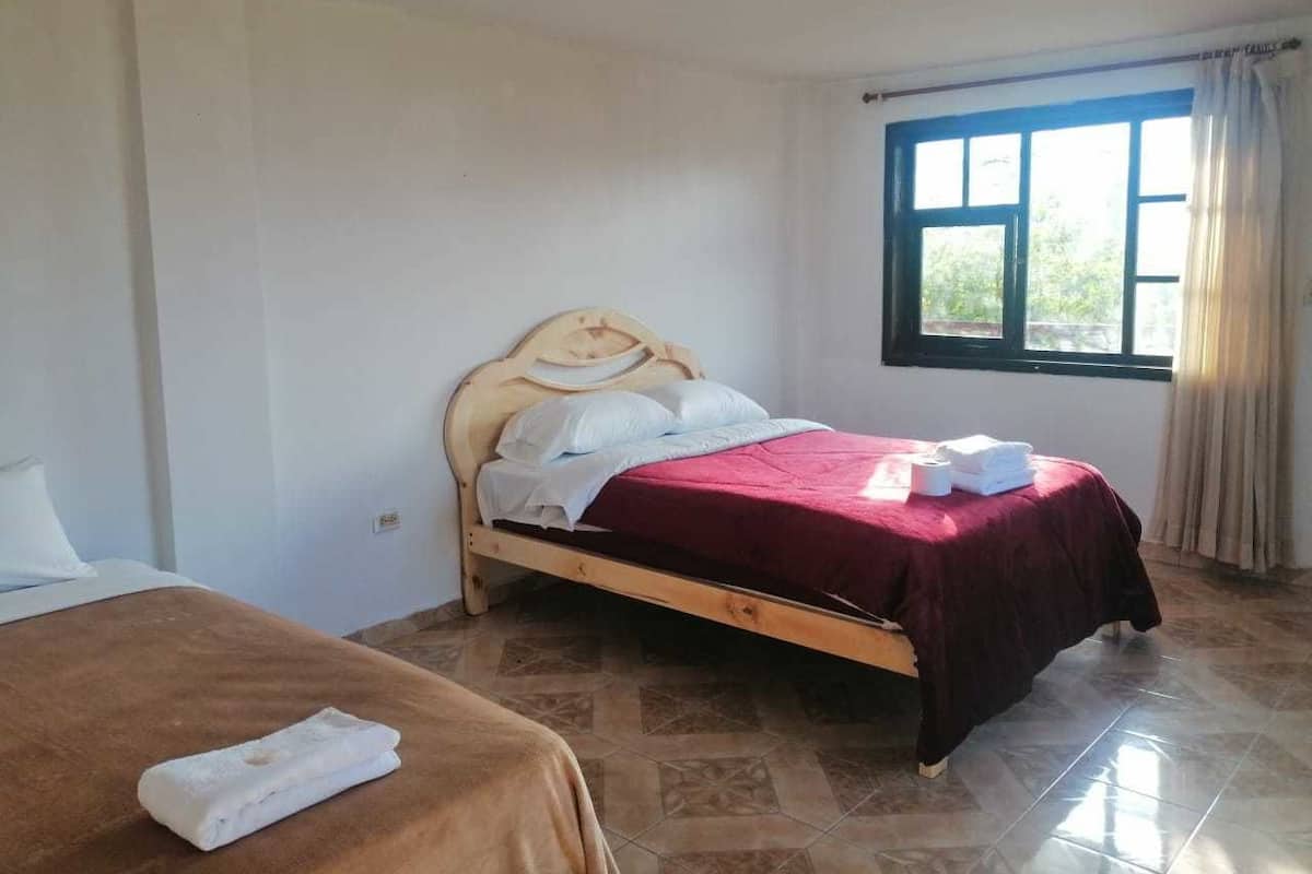 A single bed with a wooden frame and a red bedspread, a bedside table with a white towel, and a window with curtains.