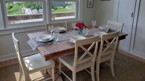 A dining room with a wooden table set for four, white chairs, and a view of a garden through large windows. - Gorgeous Views of Harbor Entrance! Walk to town, east side location. (Boothbay Harbor)