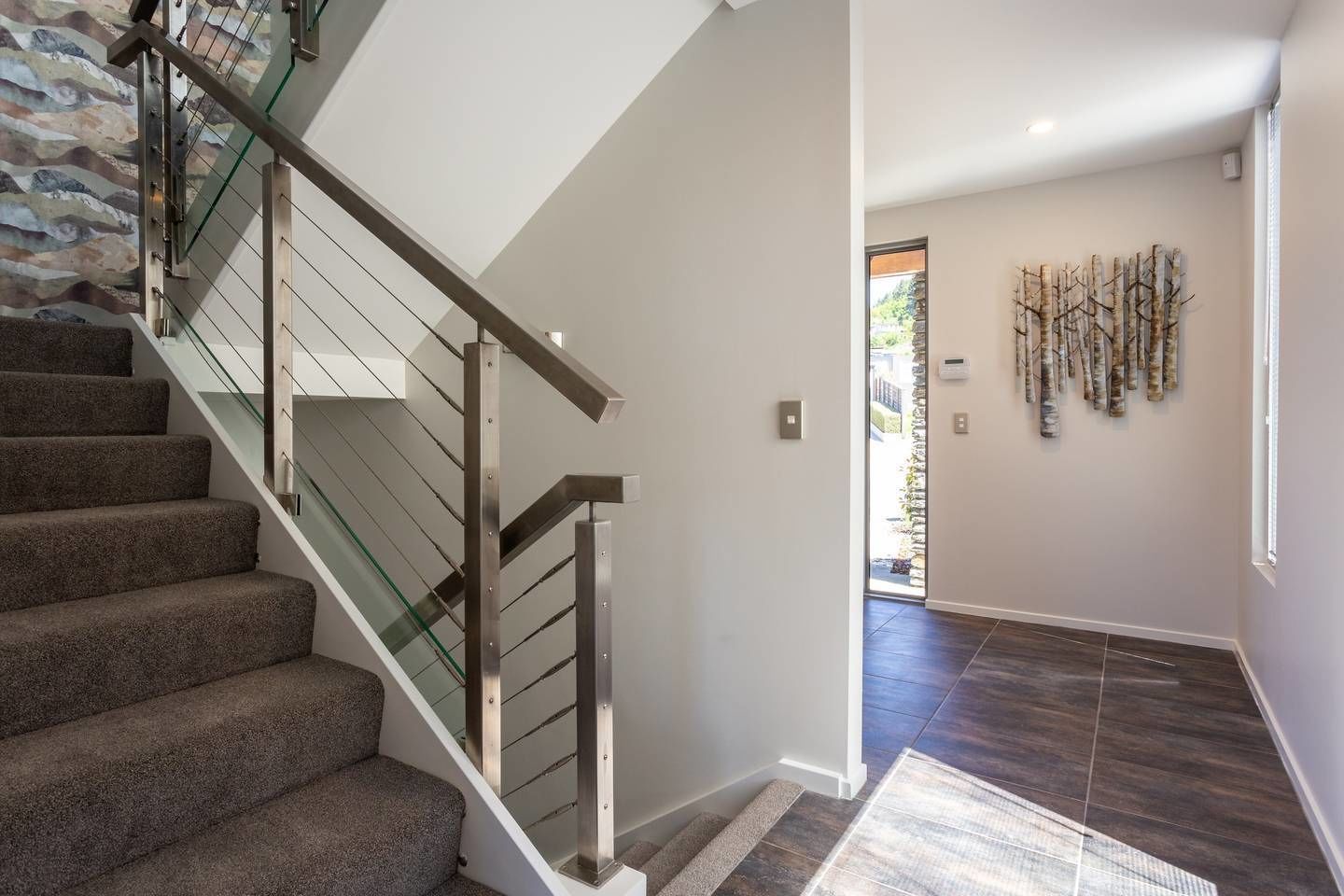 A modern staircase with glass railings and carpeted steps leading to a hallway with a dark wood floor and a decorative wall piece.