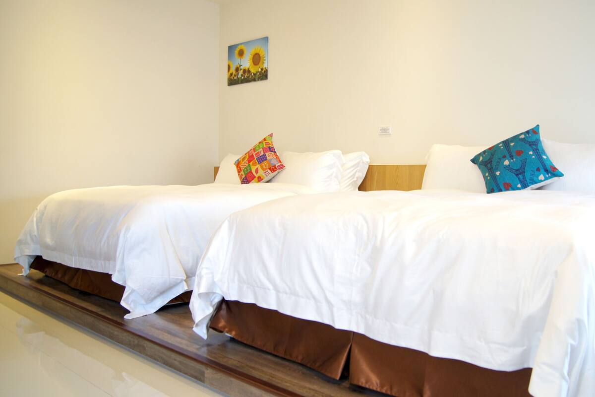 A hotel room with two beds, each with white bedding and brown headboards. There is a colorful pillow on one bed and a blue patterned pillow on the other. A painting of sunflowers hangs on the wall above the beds.