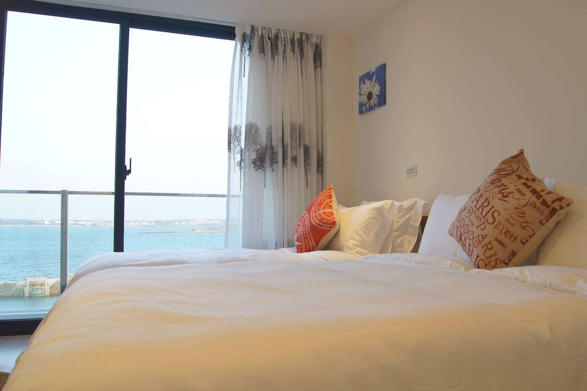 A hotel room with a large bed, a view of the sea, and a window with sheer curtains.