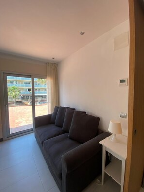 Ein Zimmer mit einem dunkelbraunen Sofa, einem weißen Nachttisch mit Lampe, einer Schiebeglastür, die zu einem Außenbereich führt, und einem an der Wand befestigten Thermostat.