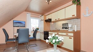 Une cuisine moderne avec des meubles en bois, un four encastré et une machine à café. On y trouve un coin repas avec une table et des chaises, ainsi qu’un téléviseur fixé au mur.