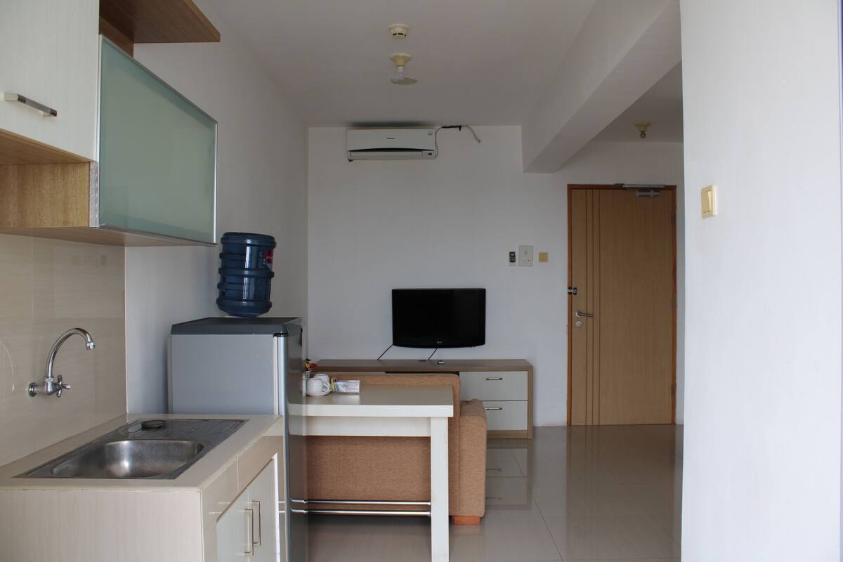 A compact kitchen with a sink, a water dispenser, a flat-screen TV, and a wooden door in the background.