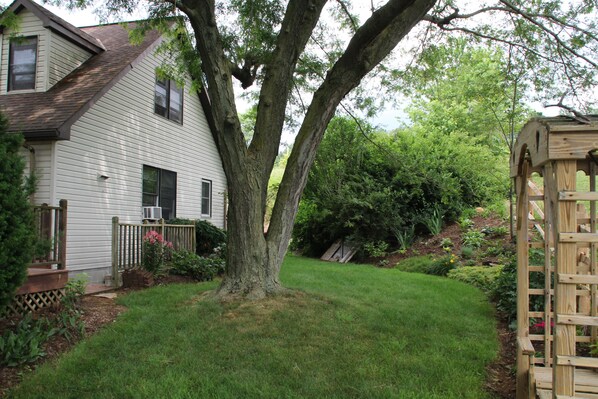 A house with a large tree in the front yard, a wooden deck, and a garden with various plants.