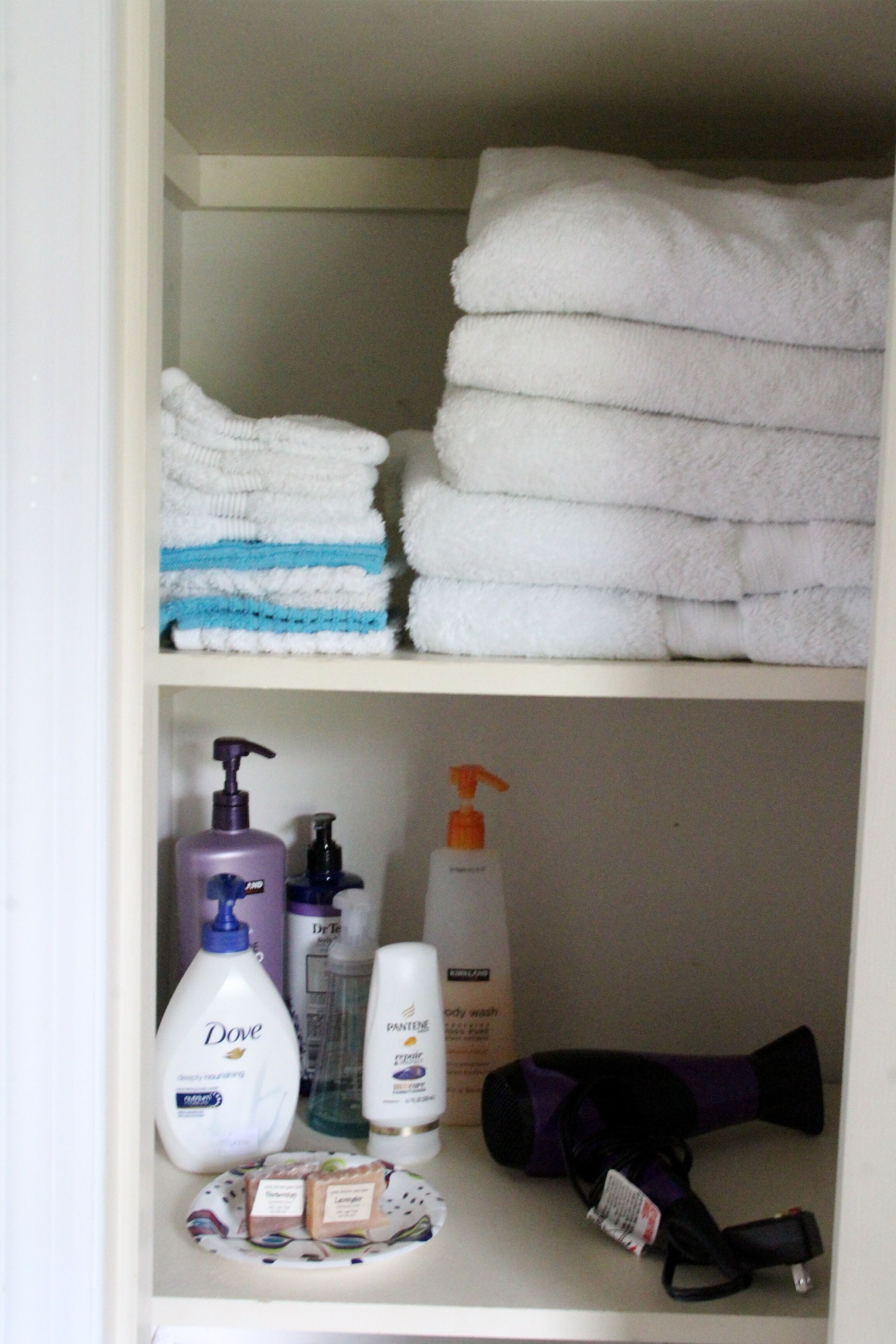Un baño con toallas blancas dobladas, jabón Dove, champú Pantene y un secador en un estante.