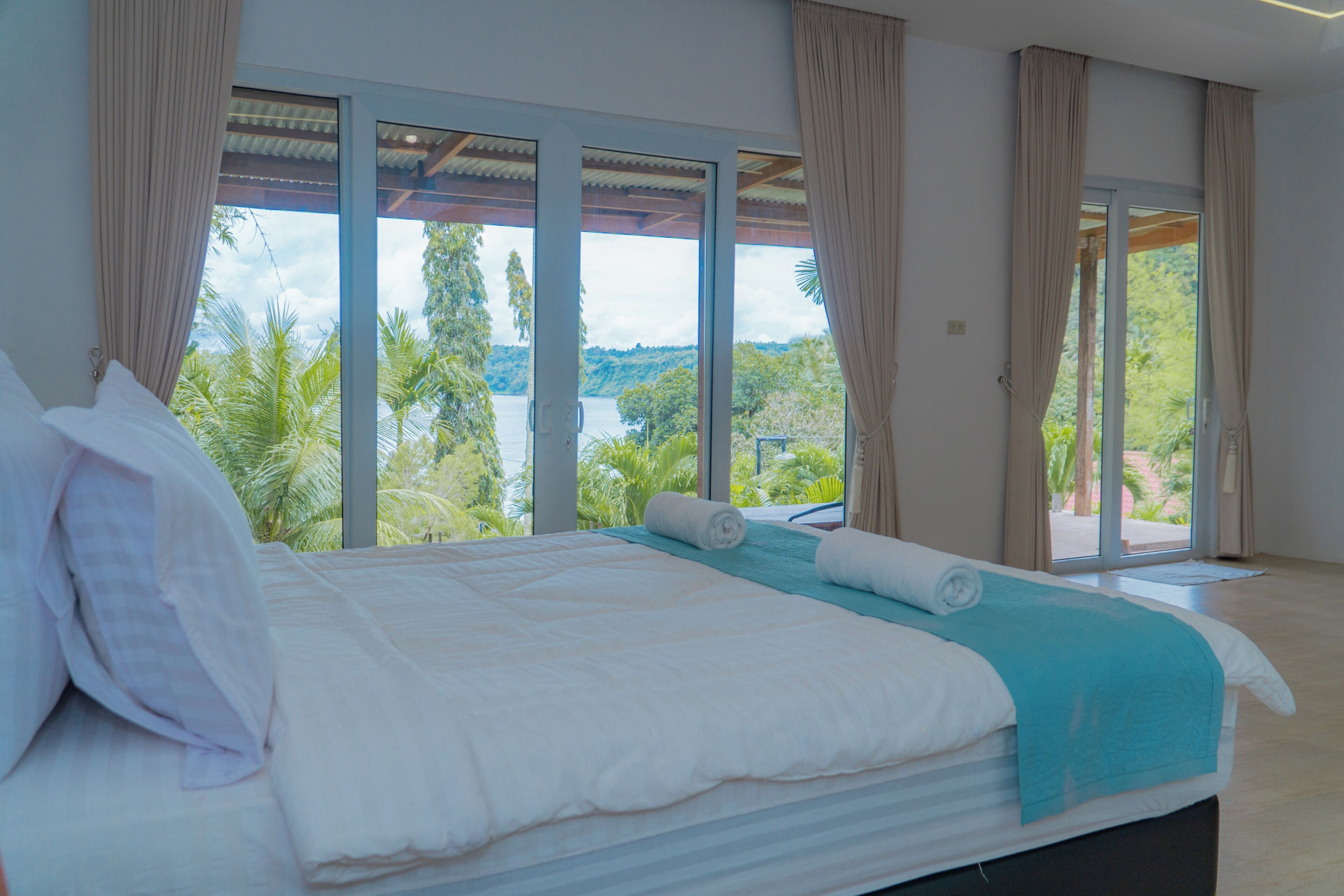 A large bed with white linens, a teal throw blanket, and a view of a tropical landscape through large windows.