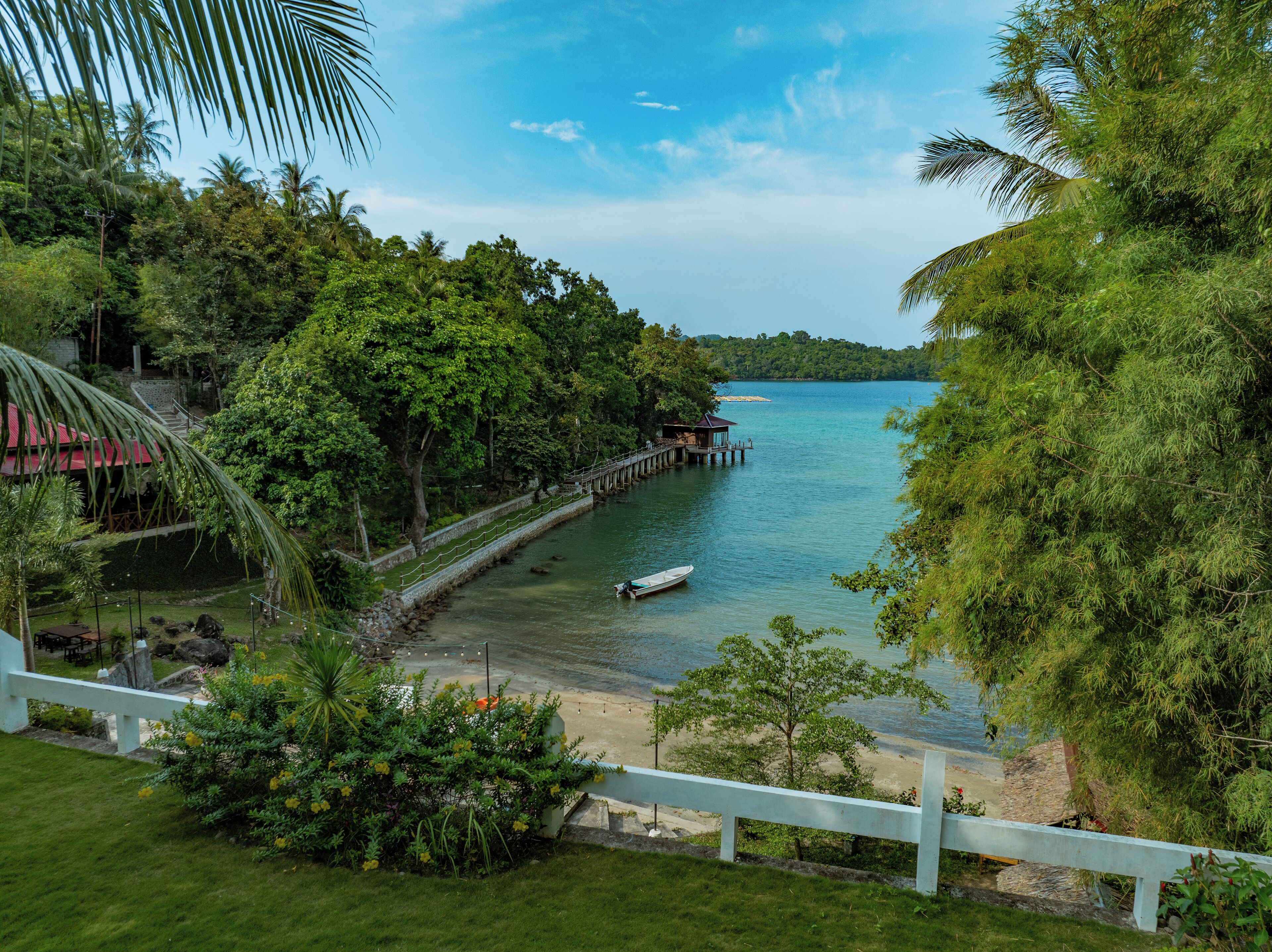 A coastal view with a wooden pier extending into calm waters, surrounded by lush greenery and a clear sky.