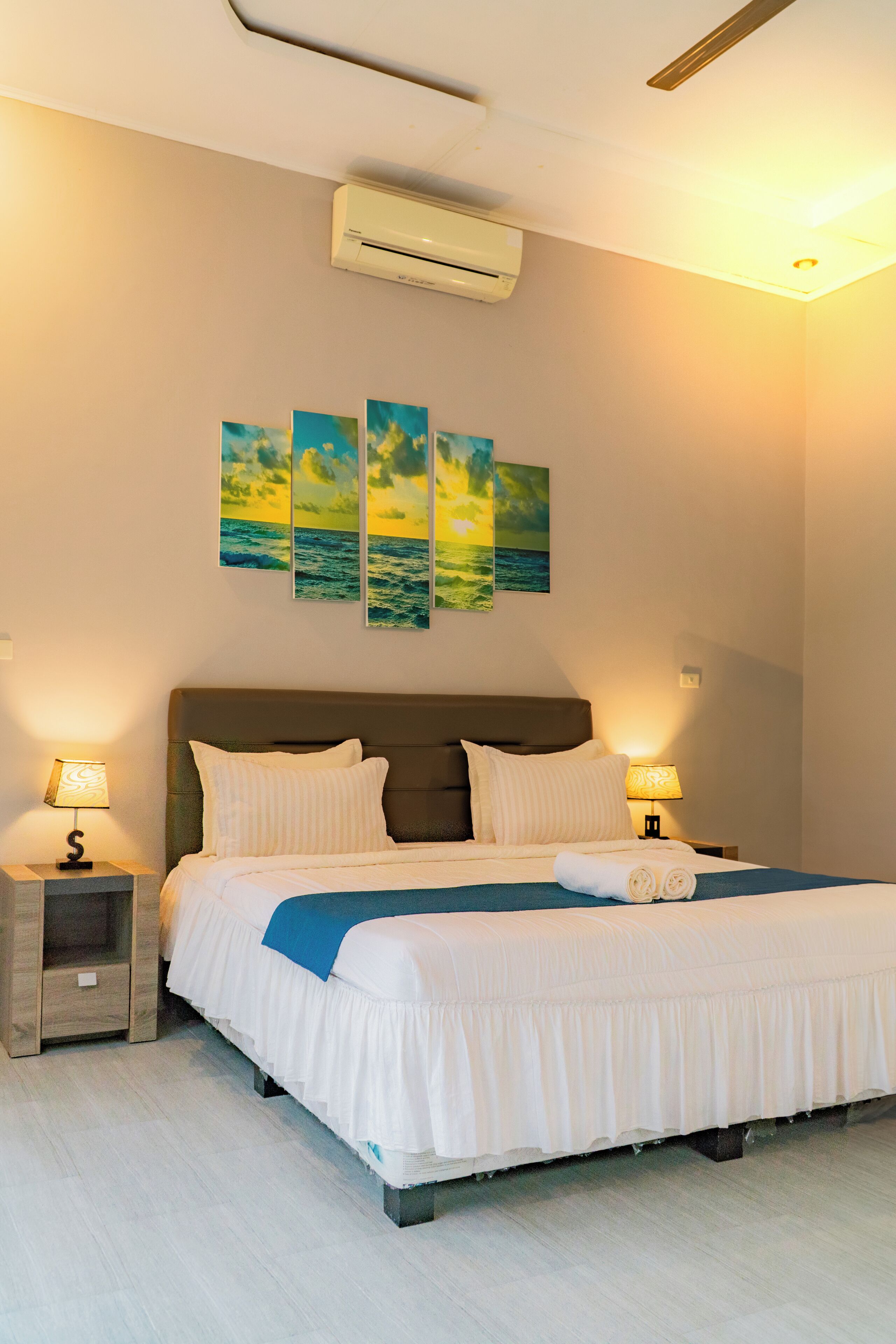 A neatly made bed with white bedding and a blue accent, a headboard, and a wall-mounted air conditioner.