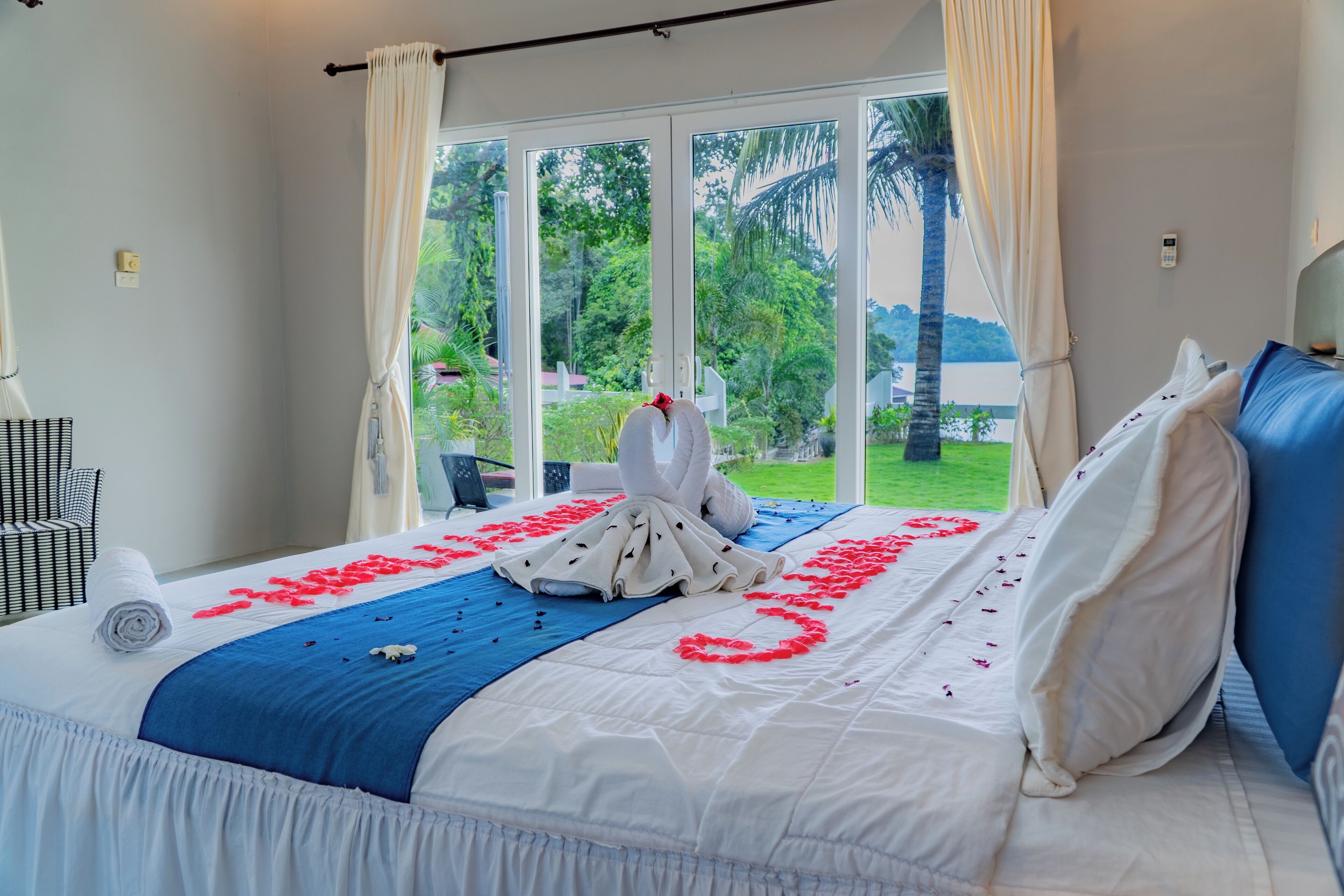 Habitación de hotel con una cama decorada con pétalos de rosa y una toalla blanca, ventanas amplias con cortinas y vistas a zonas verdes.