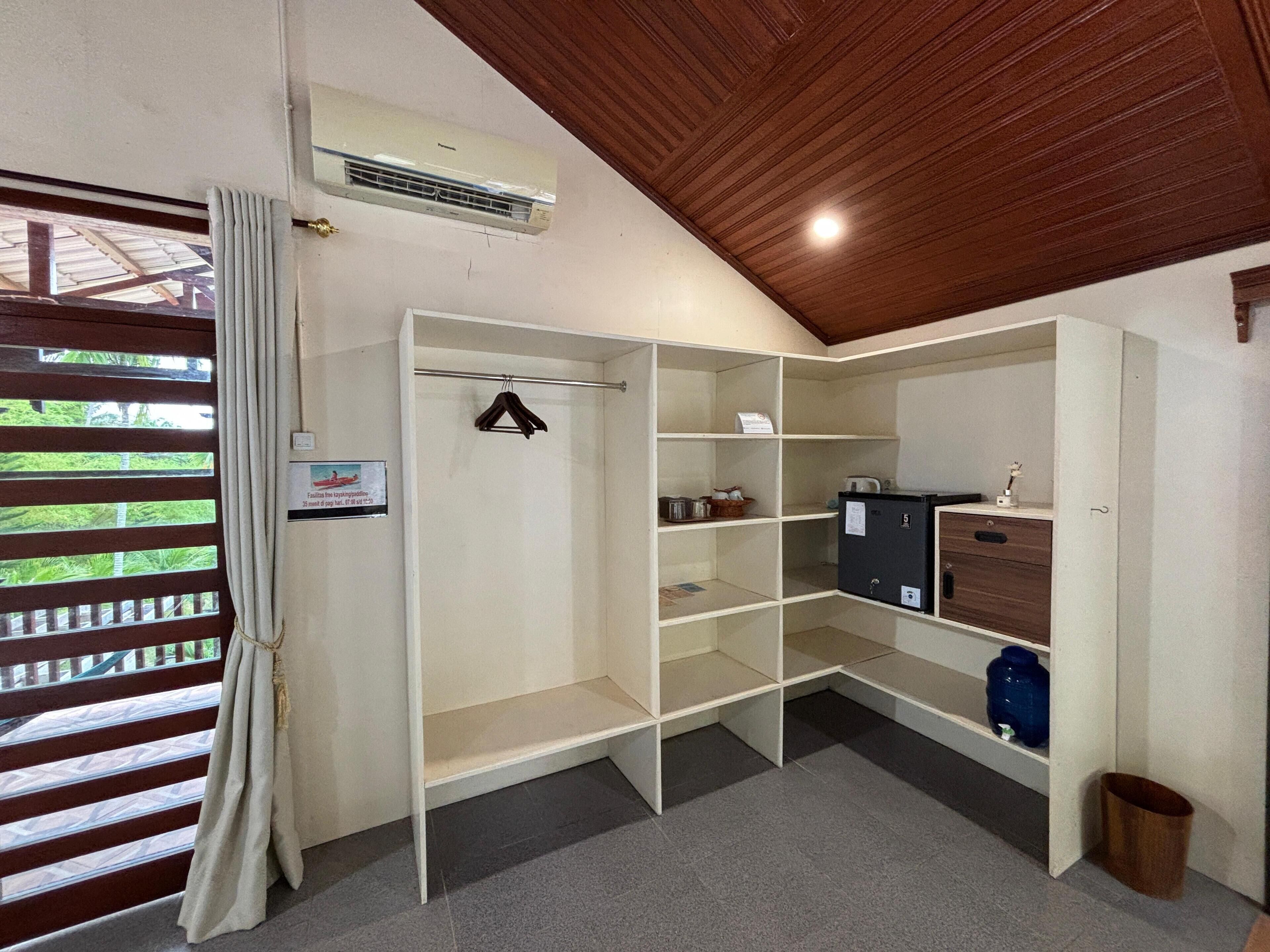 A room with a wooden ceiling, a white wardrobe, a small cabinet, and a trash bin.