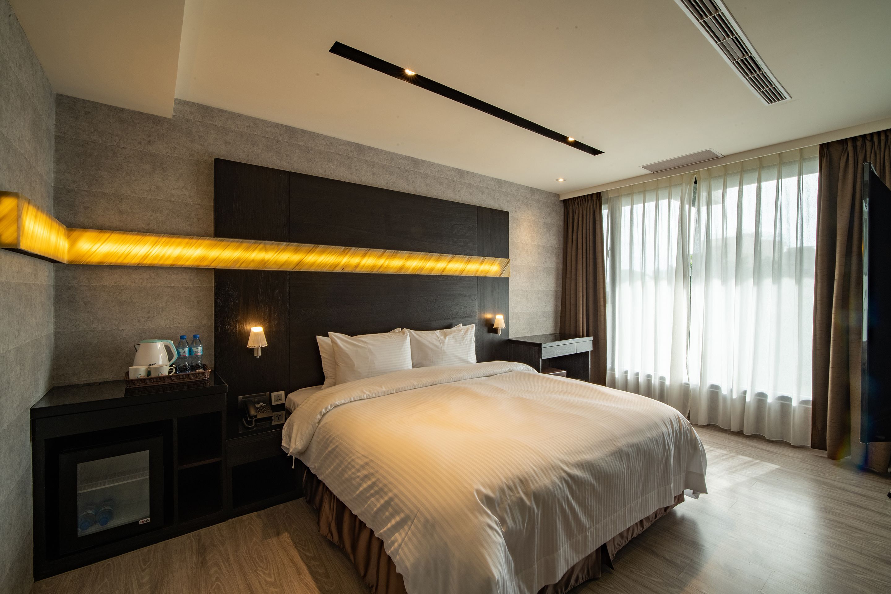 A modern hotel room with a large bed, bedside tables, a built-in TV, and a large window with curtains.