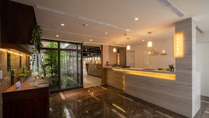 Un lobby moderno de hotel con un mostrador de recepción, una puerta de cristal que da a un jardín y un letrero con la palabra 'HELLO'.