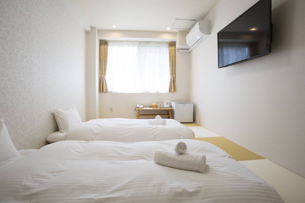 A hotel room with two beds, a TV mounted on the wall, a desk with a computer, and a window with curtains.