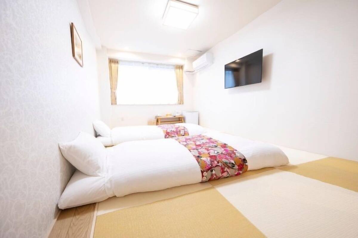 A hotel room with a bed, a television, a window, and a small bedside table.