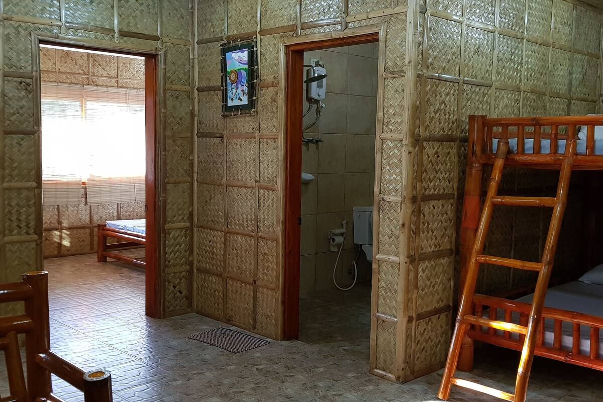 A room with a wooden bunk bed, a wooden chair, a shower area, and a bed with a blue mattress.