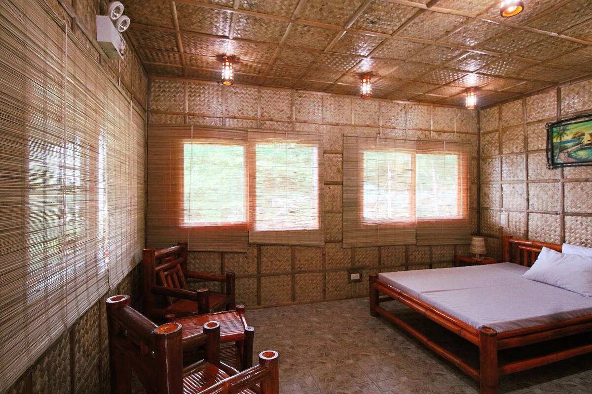 A room with a bed, a chair, and a table. There are three windows with blinds.