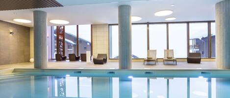 En moderne indendørs swimmingpool med klart, blåt vand, omgivet af liggestole og store vinduer med udsigt ud