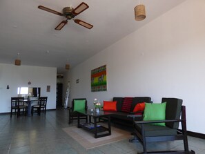 Ein geräumiges Wohnzimmer mit Deckenventilator, einem Essbereich mit Stühlen, einer Couch mit Kissen und einem Gemälde an der Wand.