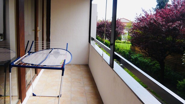 Un balcón con una silla y una mesa de metal, suelo de baldosas y vistas a un jardín con árboles y una valla.
