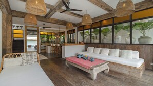 A spacious living area with a large sofa, a dining table, and a ceiling fan. There are large windows with potted plants and a view of the outdoors. - Villa Metisse by Villa Finder (Seminyak)