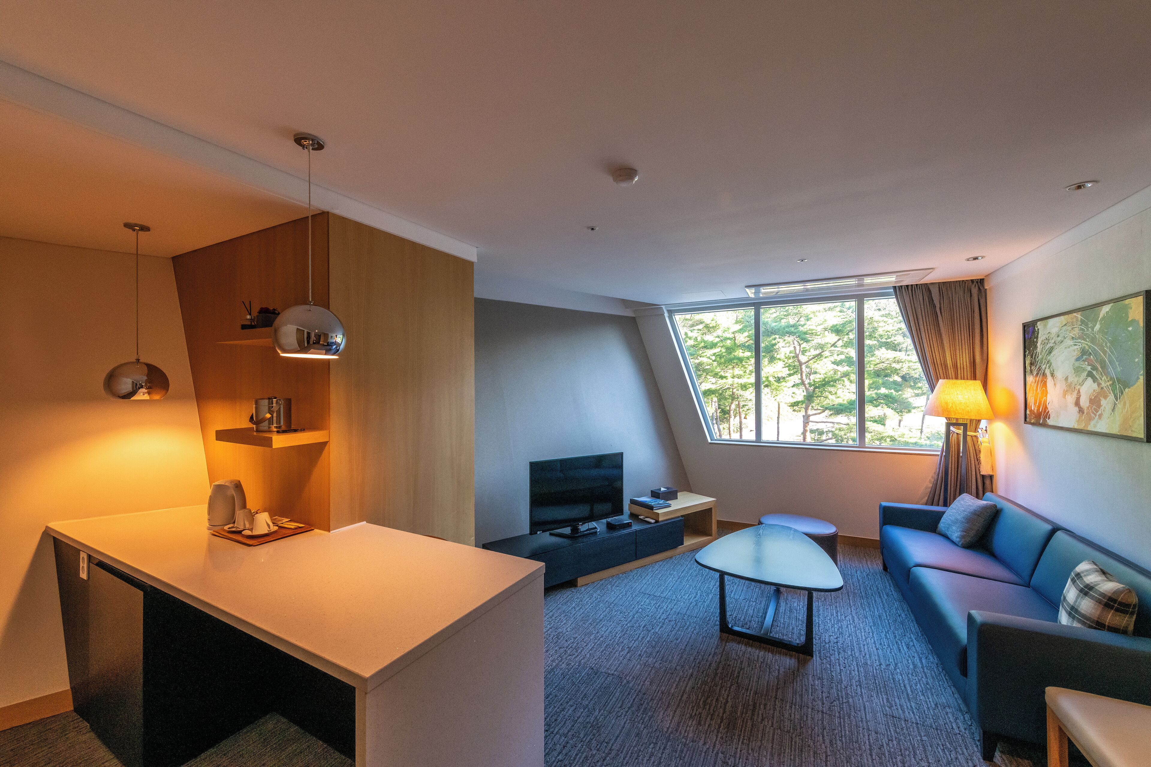 A modern hotel room with a flat-screen TV, a small dining table, a sofa, and a large window offering a view of trees.