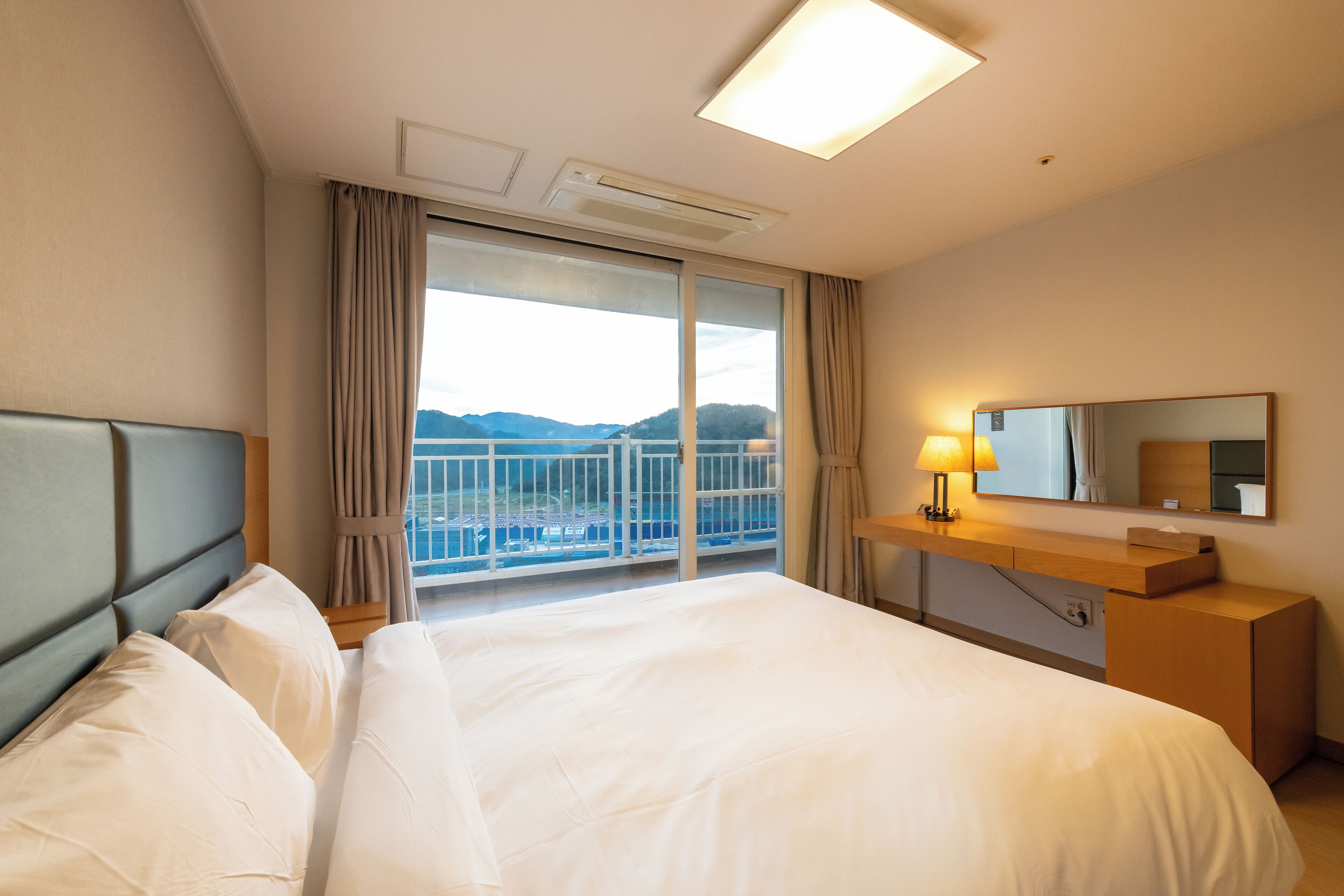 A hotel room with a large bed, a bedside table with a lamp, a mirror, and a view of the mountains through a large window.