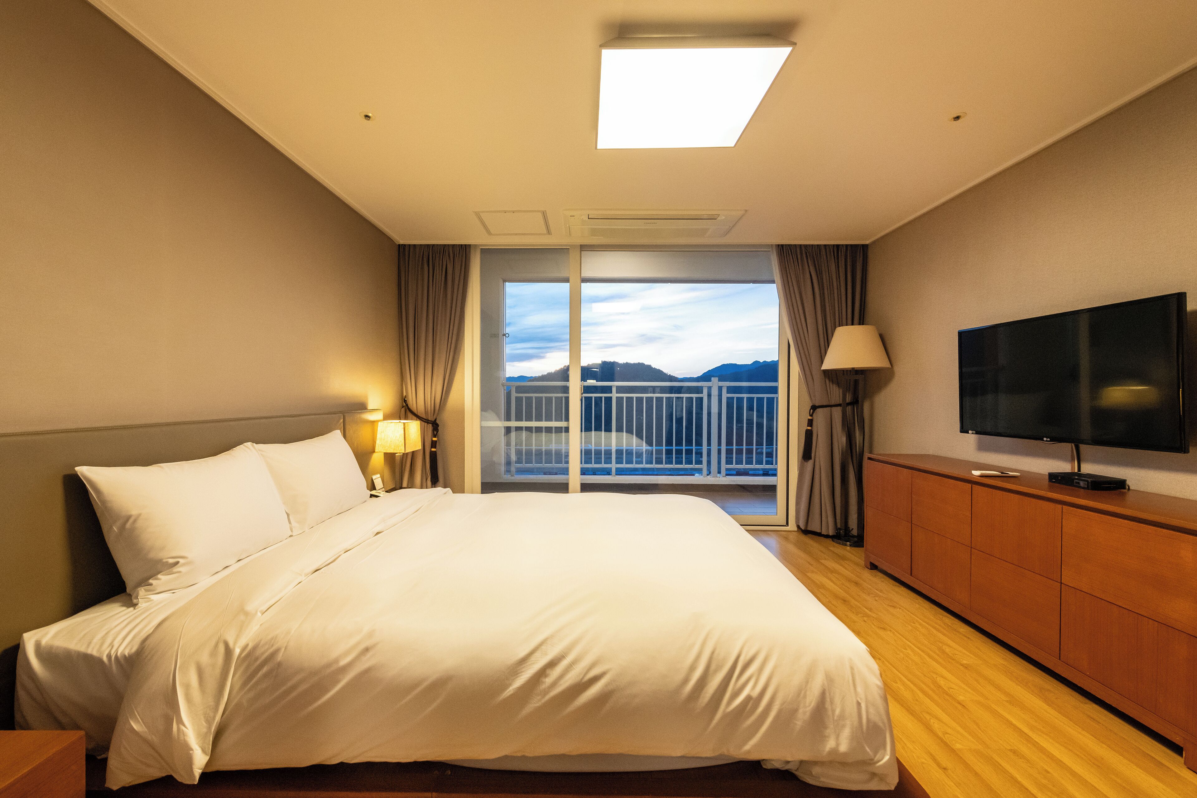 Habitación de hotel con una cama grande, televisión, cómoda de madera y balcón con vista a las montañas.