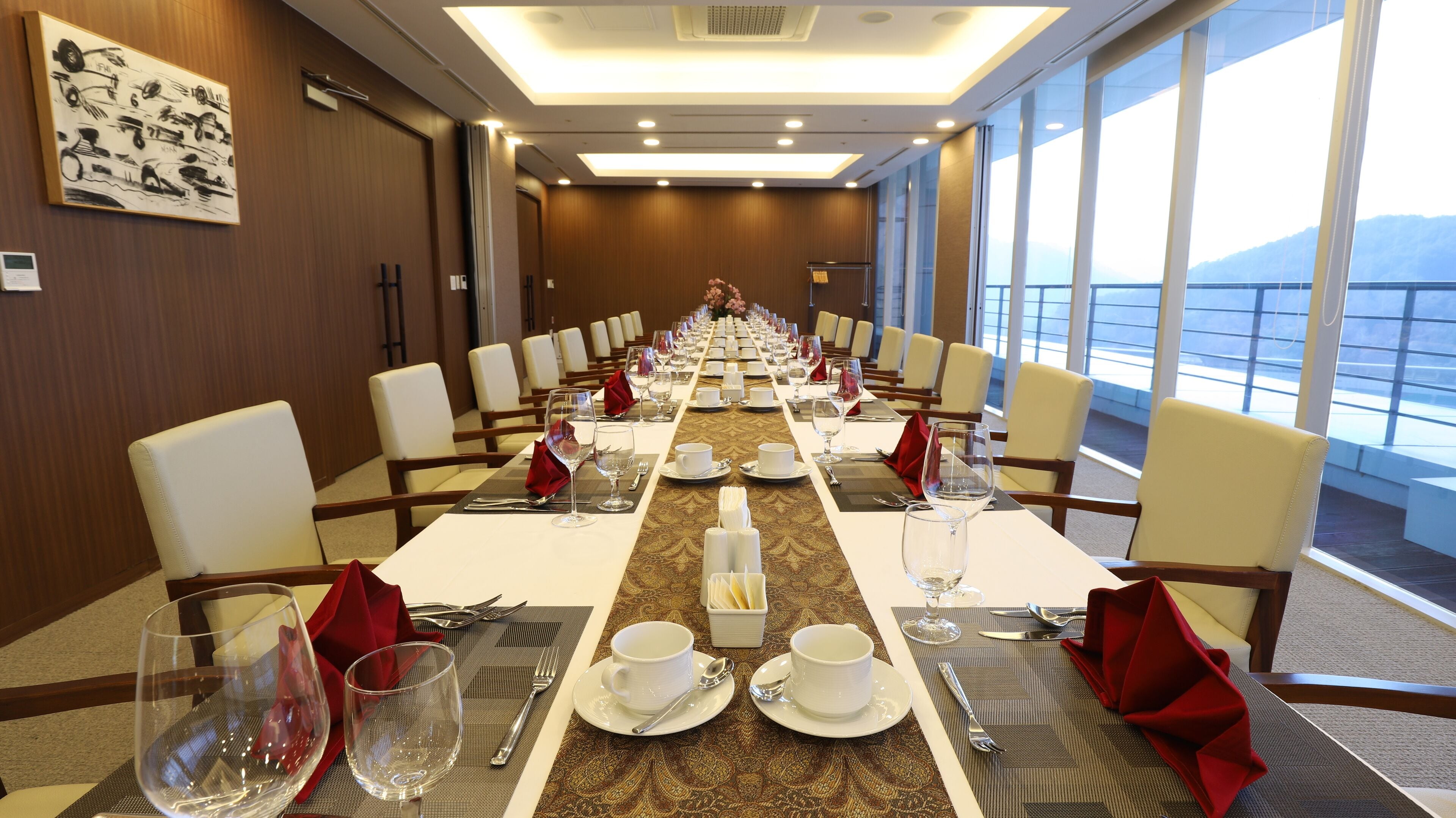 Un salón de conferencias con una larga mesa dispuesta para una reunión, ventanas amplias con vistas y un cuadro en la pared.