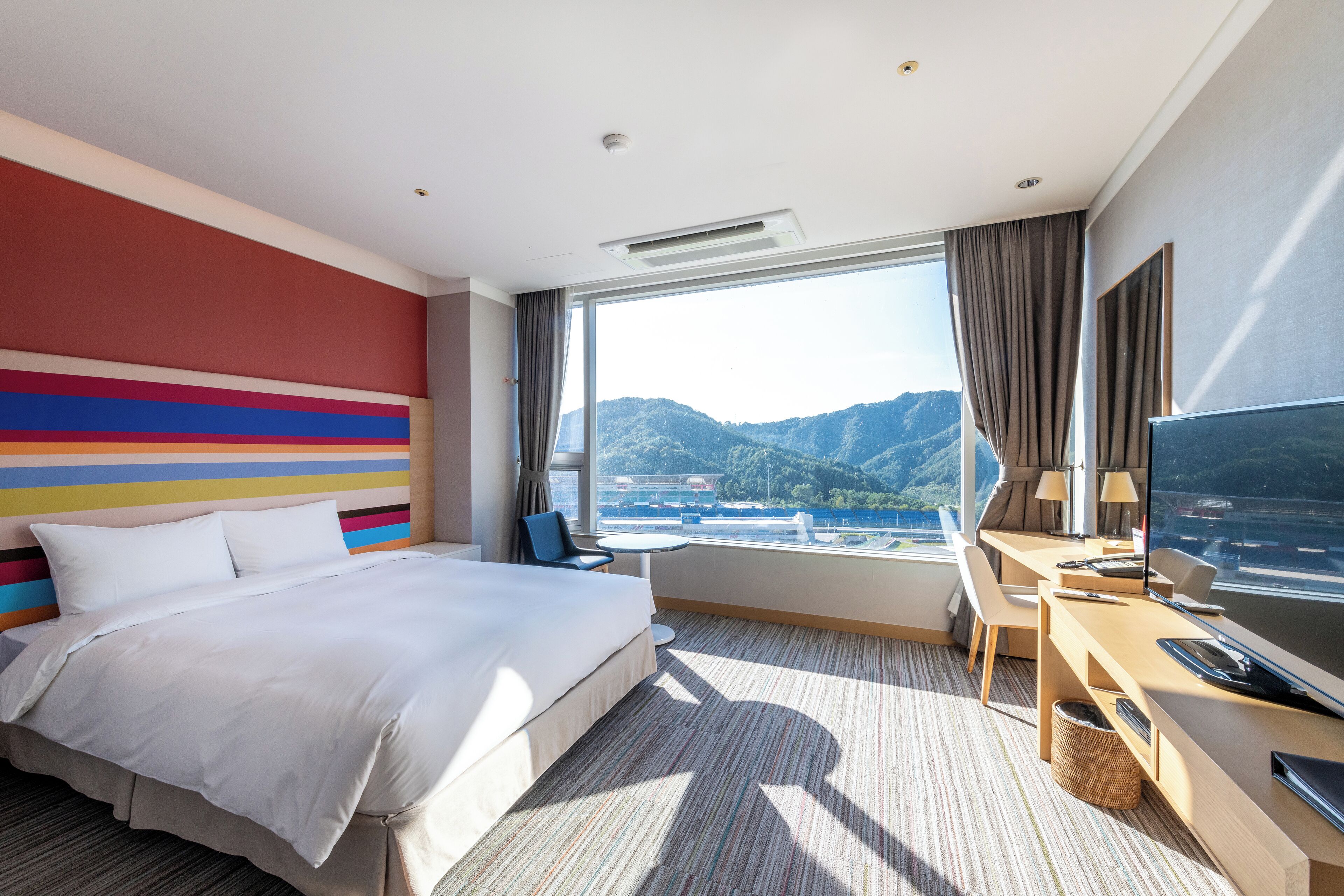 A hotel room with a large bed, a desk with a TV, and a view of mountains through the window.