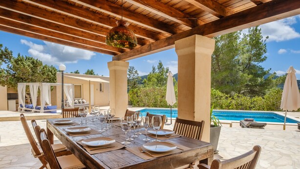 An outdoor dining area with a wooden table set for six, surrounded by chairs, with a pool and greenery in the background. - Fantastic Villa with Pool, Wi-Fi, Terrace, Garden and Sea View (Sant Josep de sa Talaia)