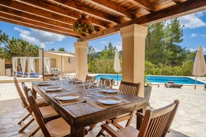 Area pranzo all'aperto con un tavolo di legno apparecchiato per sei persone, circondato da sedie, con una piscina e vegetazione sullo sfondo.