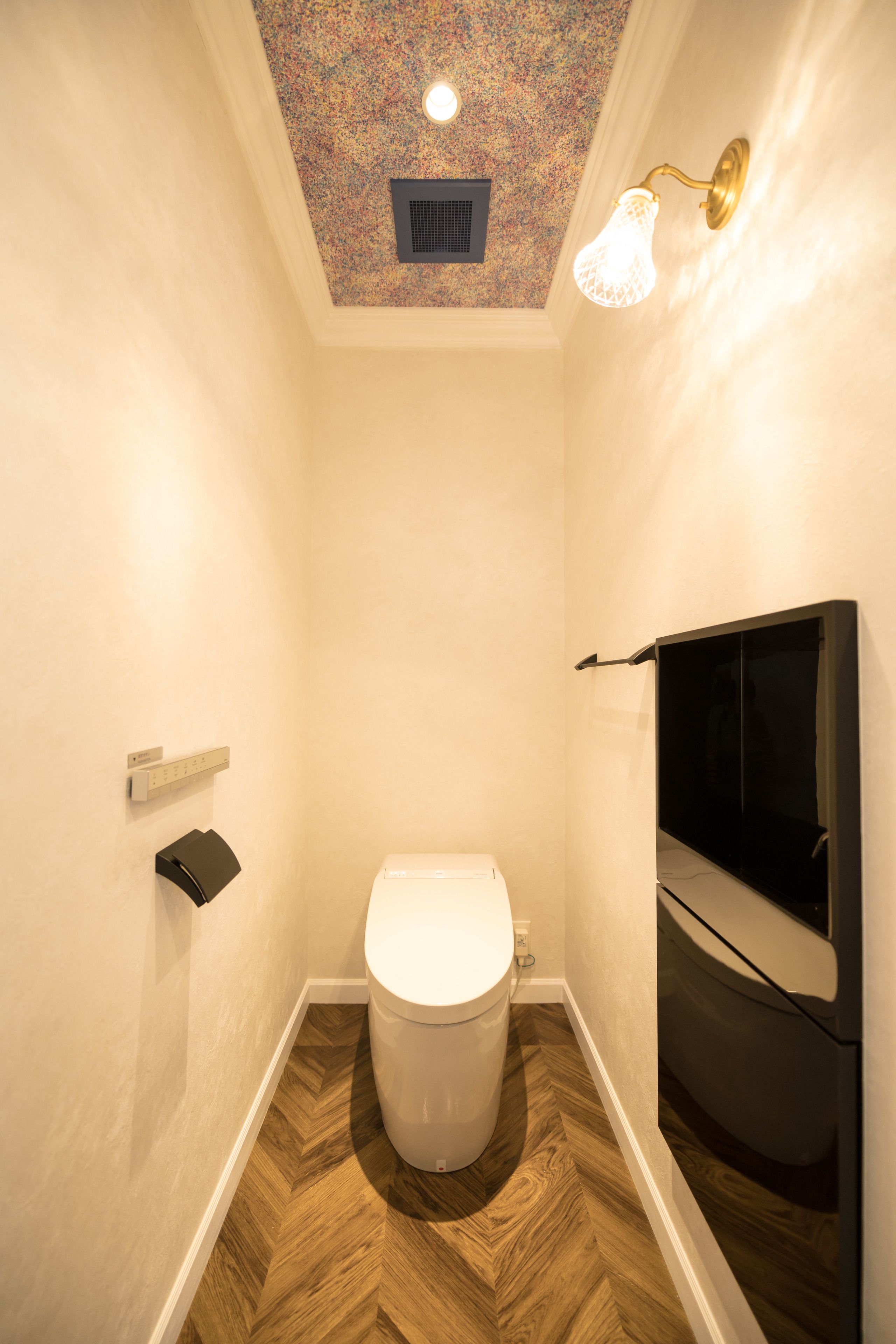 A small bathroom with a toilet, a wooden floor, a ceiling with a decorative pattern, and a wall-mounted light fixture.