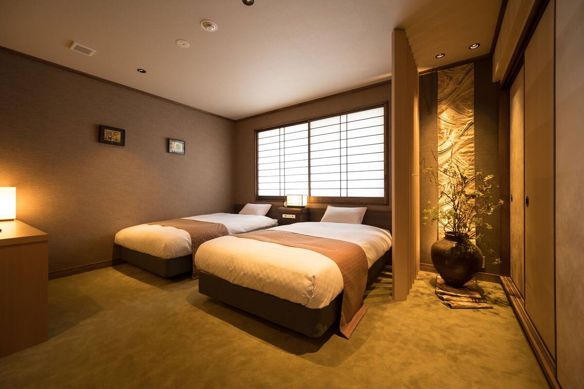 Hotelkamer met twee bedden, een groot raam met zonwering, een nachtkastje met een lamp en een grote vaas met planten.