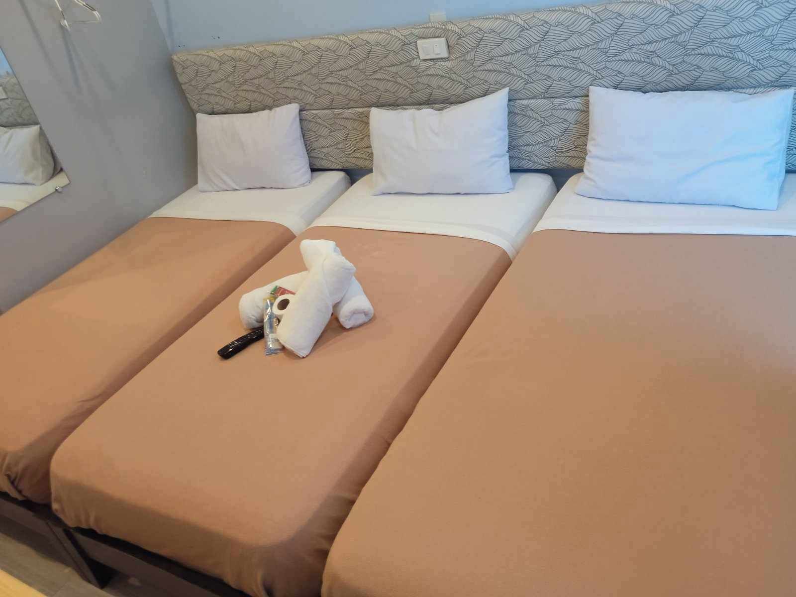 Habitación de hotel con tres camas, cada una con una colcha marrón y almohadas blancas. Un oso de peluche blanco está colocado en la cama central.