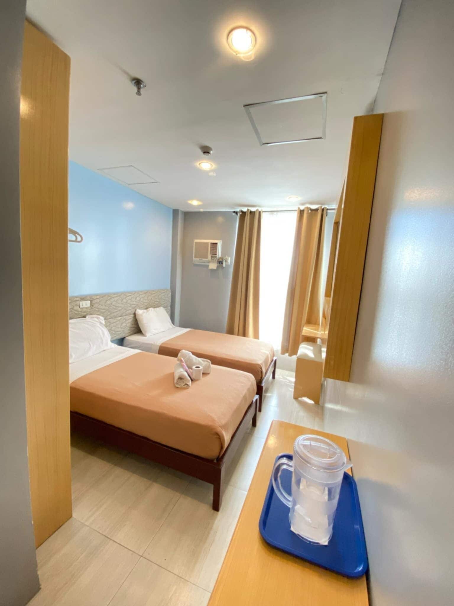A hotel room with two beds, a wooden nightstand, a bathroom mirror, and a window with curtains.