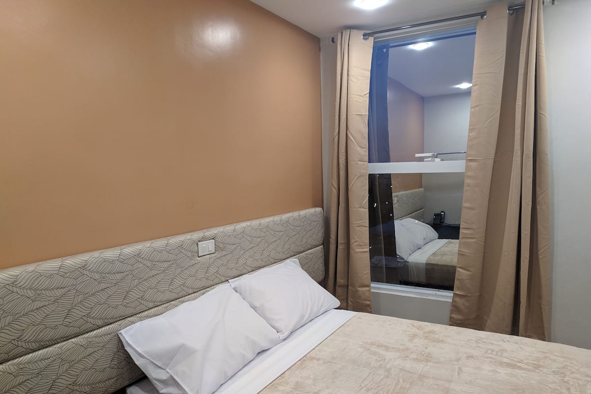 Ein Hotelzimmer mit Bett, Kopfteil, einem Fenster mit Vorhängen und einem sichtbaren Badezimmerbereich.