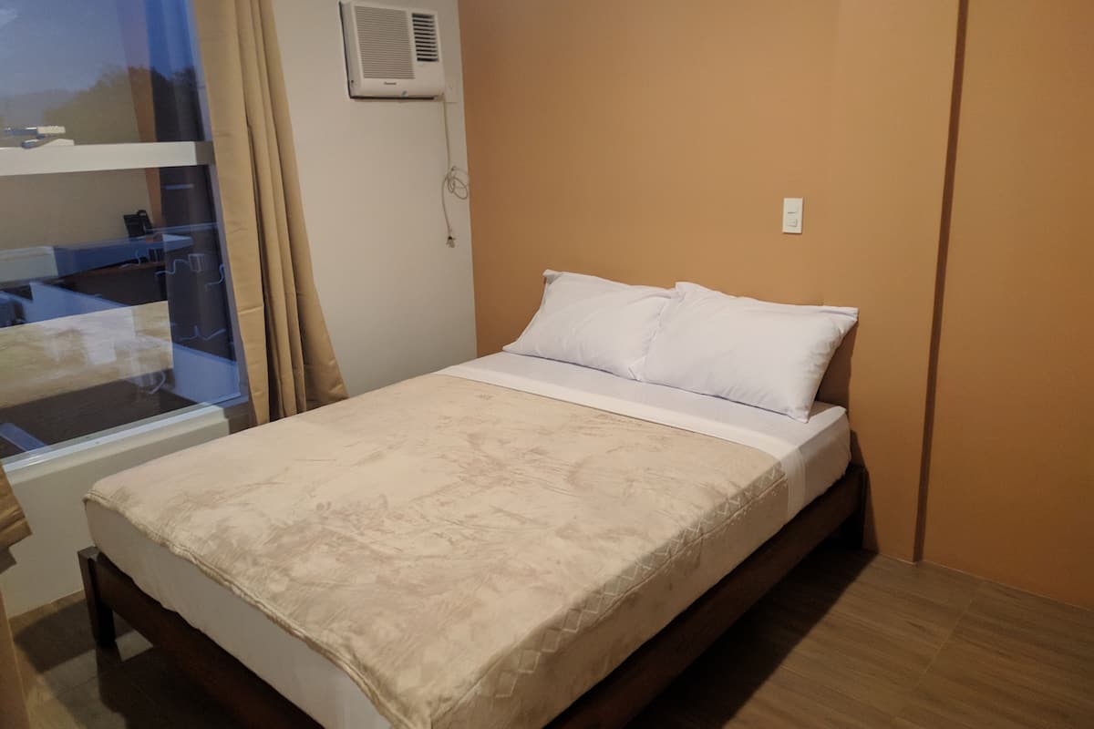 Ein ordentlich bezogenes Bett mit weißer Decke und Kissen, ein Holz-Kopfteil, ein Fenster mit Vorhängen und ein Klimagerät an der Wand.