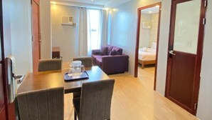 Habitación de hotel con una mesa de comedor de madera, dos sillas, un sofá morado, una cama y una puerta con un panel de vidrio satinado.