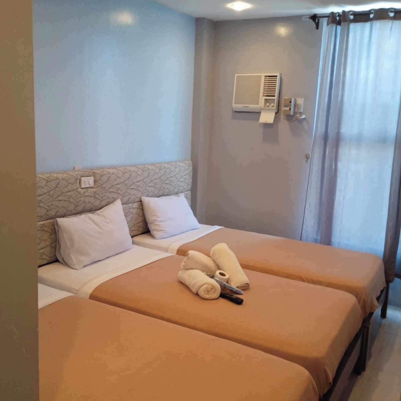 A hotel room with two beds, a ceiling fan, an air conditioning unit, and a window with curtains.