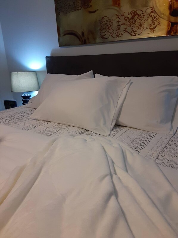 A neatly made bed with white linens and a patterned bedspread, a nightstand with a lamp and a digital clock, and a decorative wall hanging above the bed.